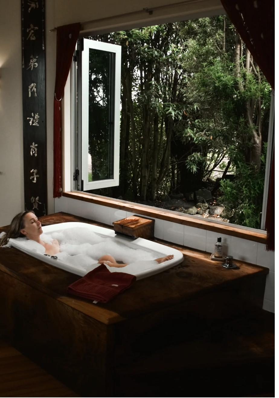 A person reclined in a conventional bath, timber and forest view through the window