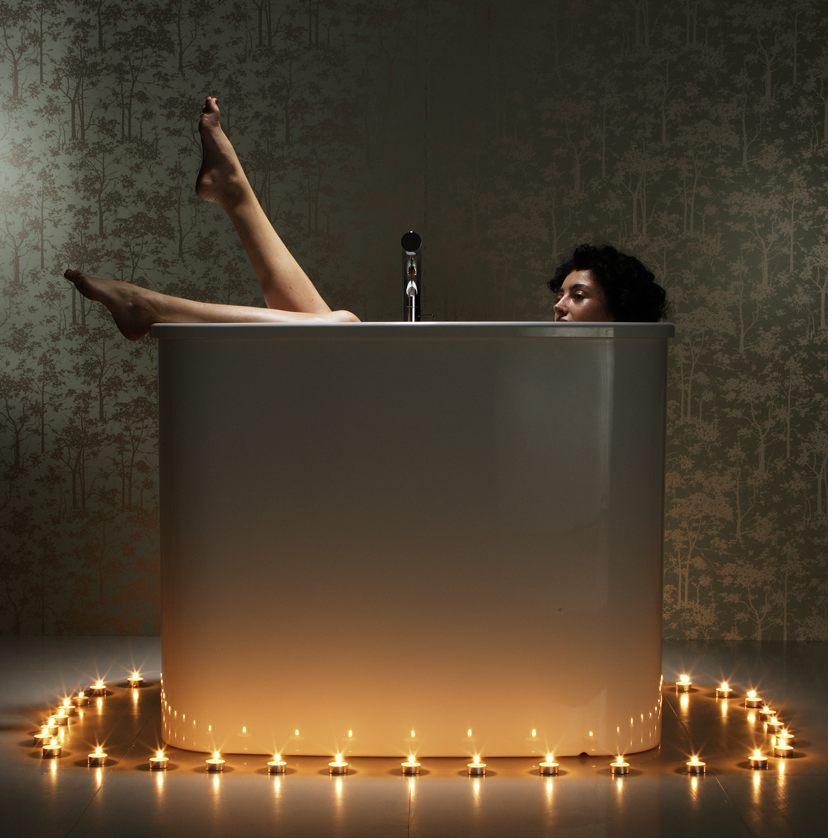 A person seated upright, deeply immersed in an AQVA deep-soak bath, candle ring in frame